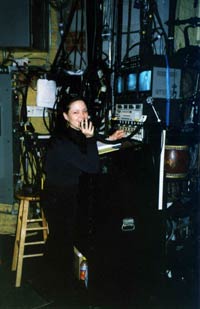About the photo: Monroe is pictured stage-managing a production of Flower Drum Song at the Virginia Theatre in New York City. Here’s her description of what you see: “I am at the ‘calling desk’ from which a stage manager runs the show. My left hand is on a cue switch for automation, and I am speaking into the paging system. Directly above the cuing switches is a panel for the stage manager’s headset contacts: Light Board Operator, Followspots, Deck, Sound, etc. On top you see three monitors. The left one is a side view of the stage (important on some shows for the safety of the performers and big moving set pieces), the middle one is a view of the whole stage, and the one on the right is a monitor of the conductor, whom I have to follow carefully. On the back of the desk you’ll see a red drum—that was one of the Flower Drums for the show.”